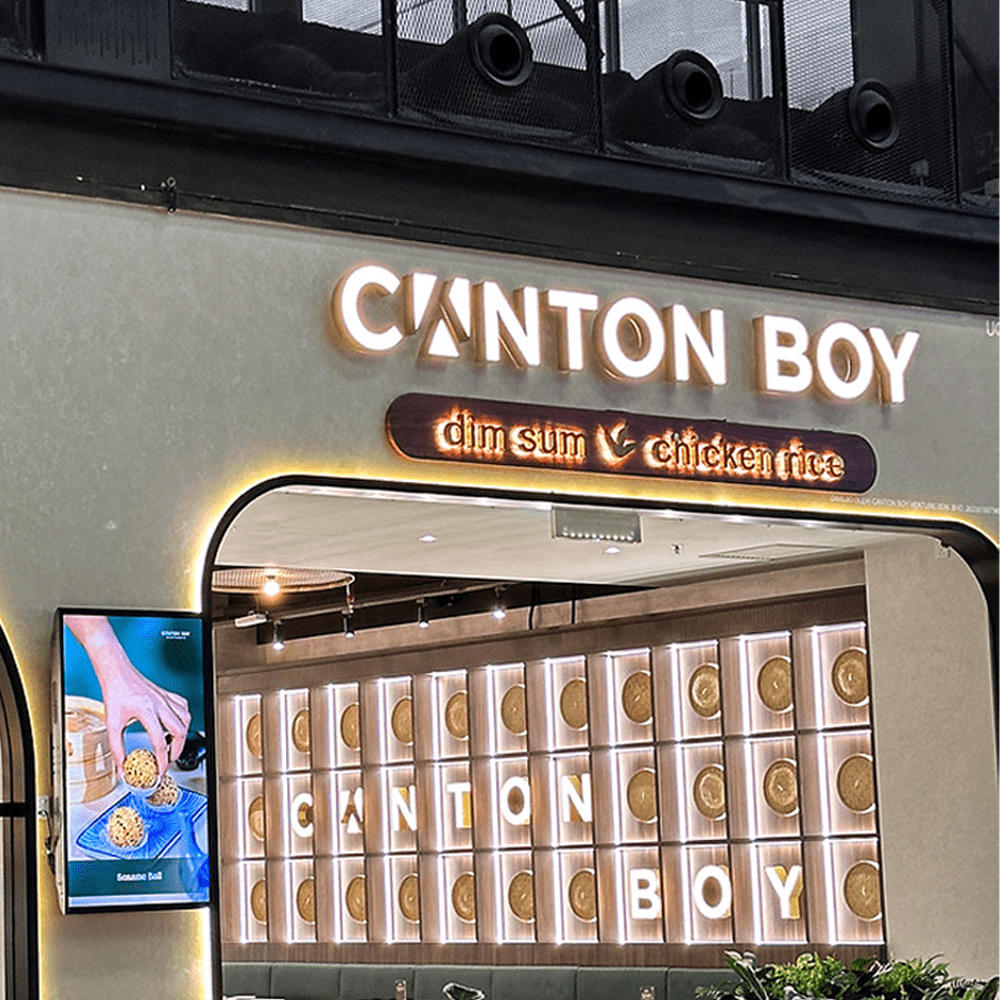 The image shows the interior entrance of a restaurant named "Canton Boy," which specializes in dim sum and chicken rice. The signage is illuminated with bold, glowing letters spelling "CANTON BOY," and a smaller sign below highlights "dim sum" and "chicken rice" with decorative lighting. The interior features a stylish, modern design with a wall panel displaying dim sum baskets and backlit letters spelling out the restaurant's name. To the left, a digital screen showcases an image of a hand holding a golden, crispy dim sum item. The atmosphere is warm and inviting, with elegant lighting and contemporary decor.